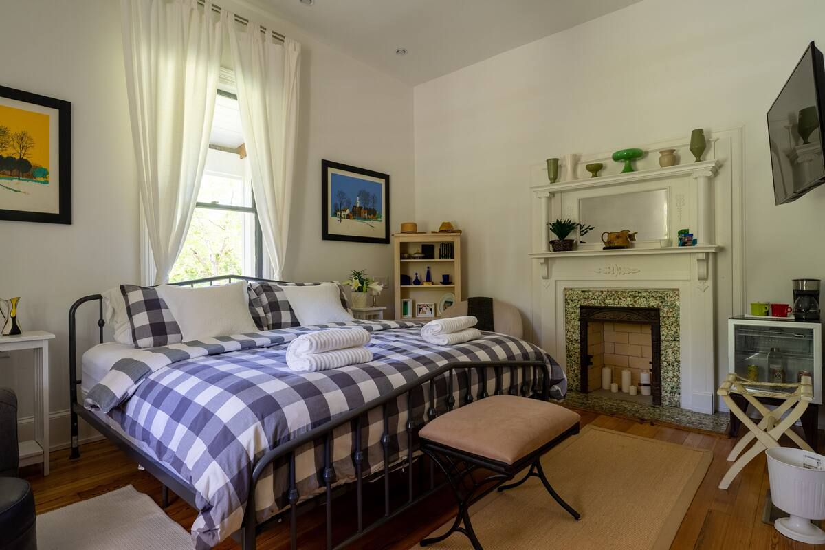 A bedroom with a checkered bedspread, a white fireplace, and a bedside table with a lamp.