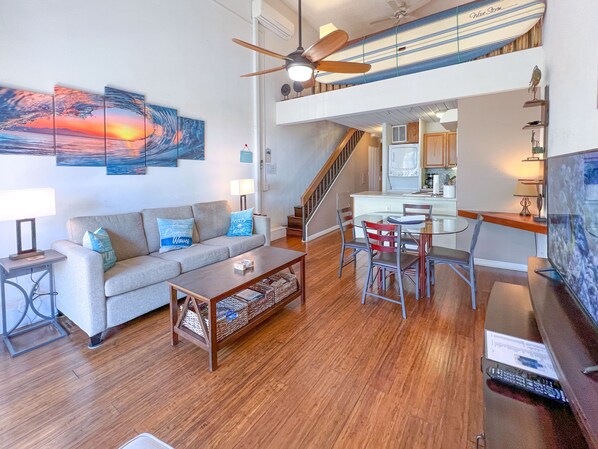 A spacious living area with a sofa, coffee table, and dining table. A ceiling fan and artwork are visible. A kitchen and staircase are in the background.