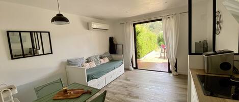 En moderne stue med sofa, spisebord med stole, et lille køkken og en skydedør til en udendørs terrasse.