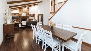 Un comedor con mesa de madera y sillas blancas, chimenea de piedra y una escalera con barandillas de madera.