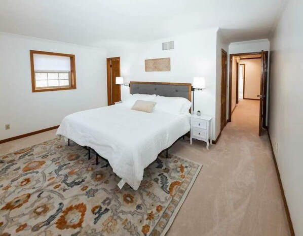 Un dormitorio con una cama grande, una mesita de noche, una lámpara de noche, una alfombra estampada, una ventana y una puerta que da a otra habitación.