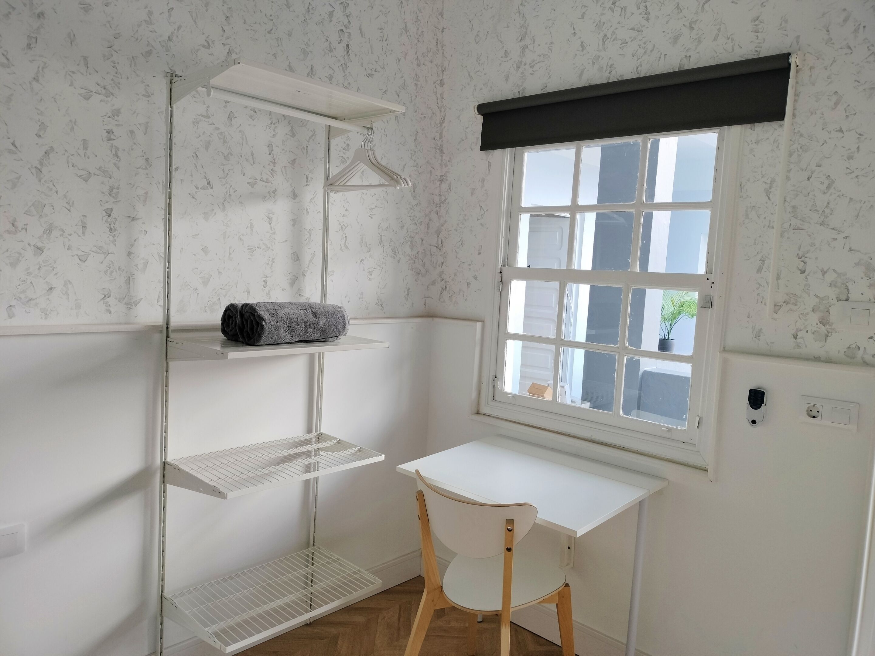 Una habitación pequeña y limpia con un escritorio blanco, una silla, un ventanal con vistas y una estantería con una toalla.