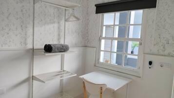 Una habitación pequeña y limpia con un escritorio blanco, una silla, un ventanal con vistas y una estantería con una toalla.