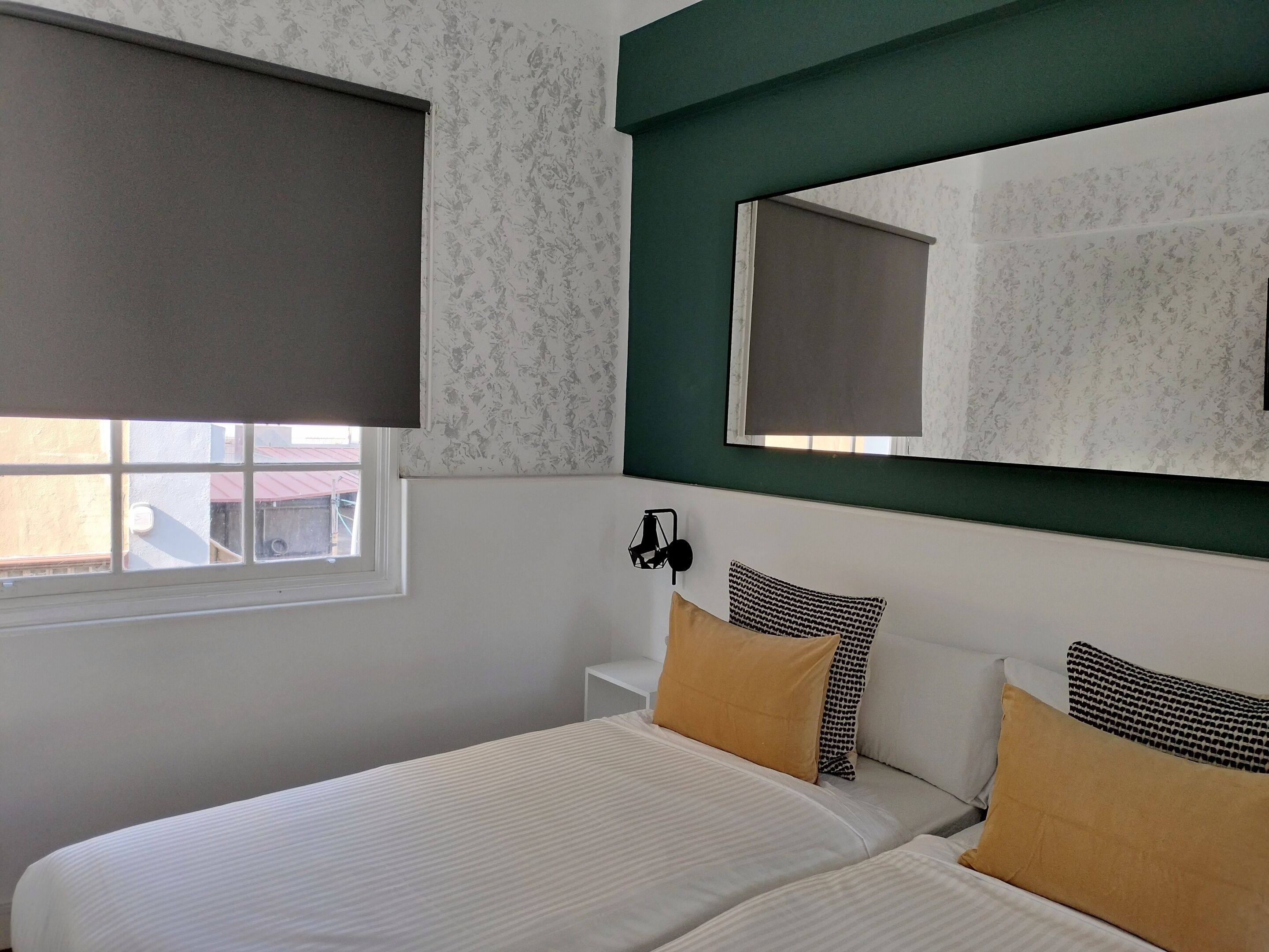 Un dormitorio con cama, dos almohadas, mesita de noche, espejo y ventana con persiana.