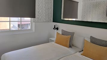 Un dormitorio con cama, dos almohadas, mesita de noche, espejo y ventana con persiana.