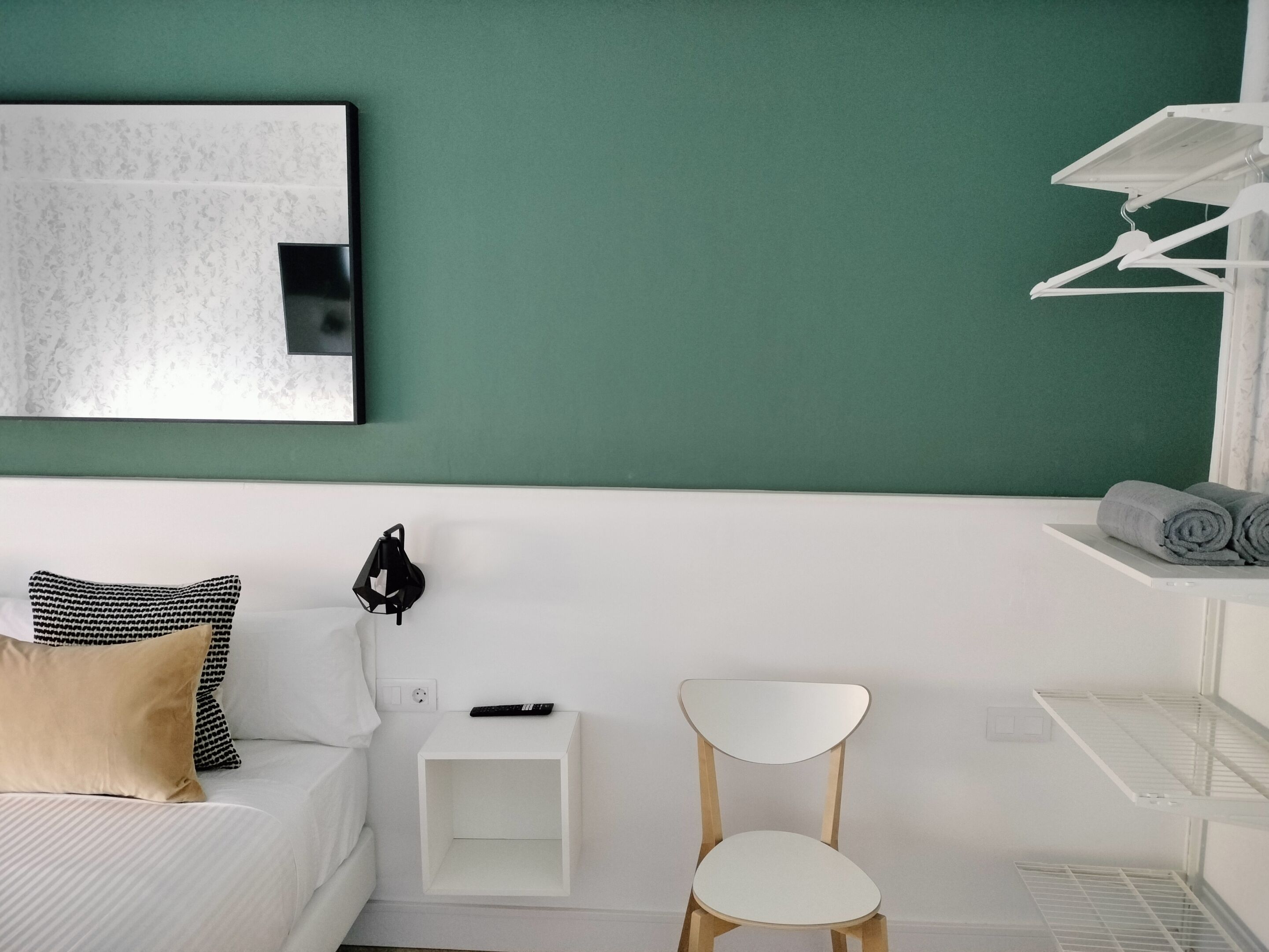 A modern bedroom with a green wall, a white bed, a white nightstand with a remote control, a white chair, and a white shelf with towels.