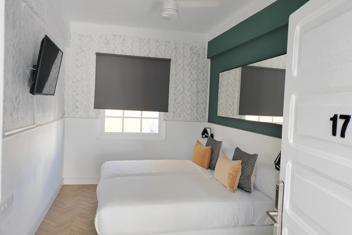 Un dormitorio con cama, televisión, ventana con persianas, puerta con número y ventilador de techo.