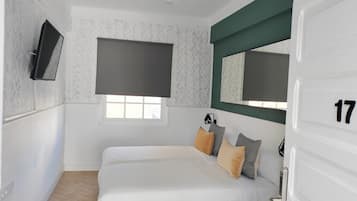 A bedroom with a bed, a TV, a window with blinds, a door with a number, and a ceiling fan.