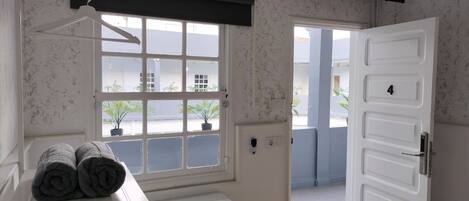 Una habitación con una mesa blanca, una ventana con plantas en macetas, una puerta blanca con un número y un ventilador de techo.