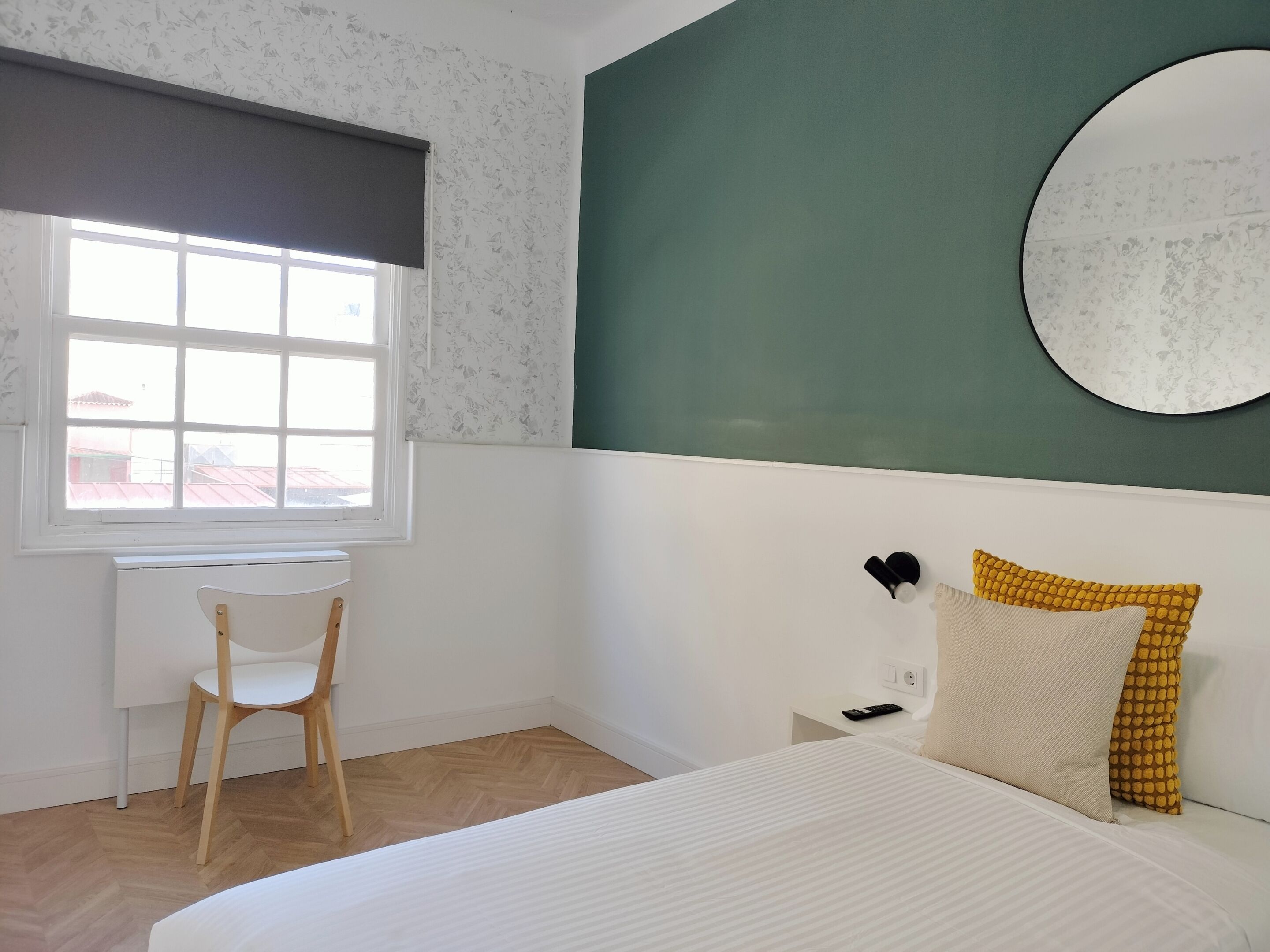 Un dormitorio con cama, mesita de noche, una silla, ventana y espejo adosado a la pared.