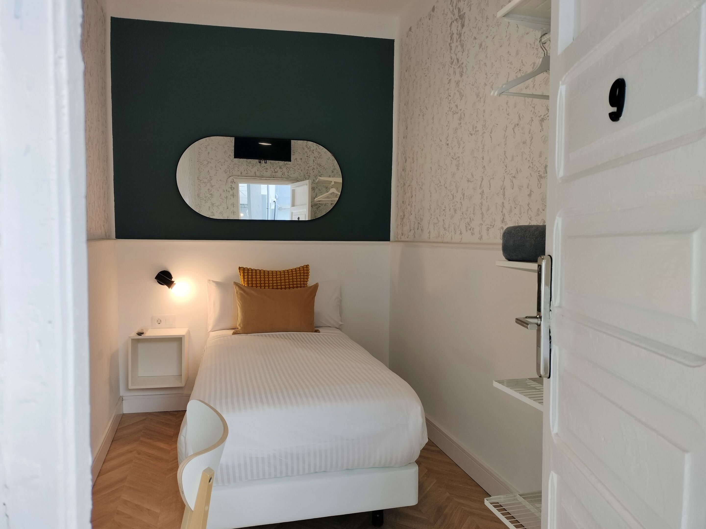 A small, neatly arranged bedroom with a bed, a nightstand, a mirror, and a door with a number.