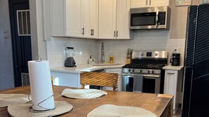 A modern kitchen with white cabinets, a wooden dining table with placemats, a stainless steel refrigerator, and a built-in oven.