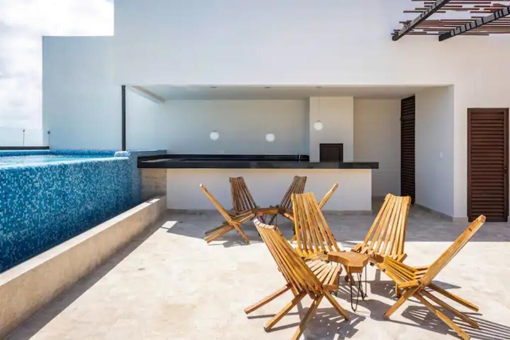 A modern outdoor seating area with wooden chairs and a table, overlooking a swimming pool and a minimalist building with a staircase.