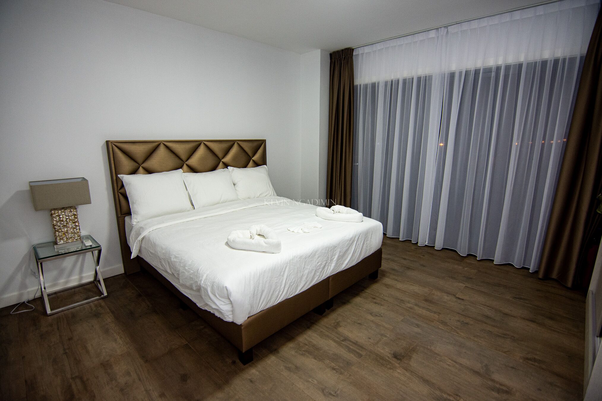 A neatly made bed with white linens and towels, a bedside table with a lamp, and a large window with sheer curtains.