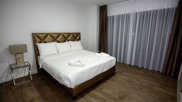 A neatly made bed with white linens and towels, a bedside table with a lamp, and a large window with sheer curtains.