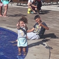 Área de piscina com uma criança na água e outra pessoa sentada perto.