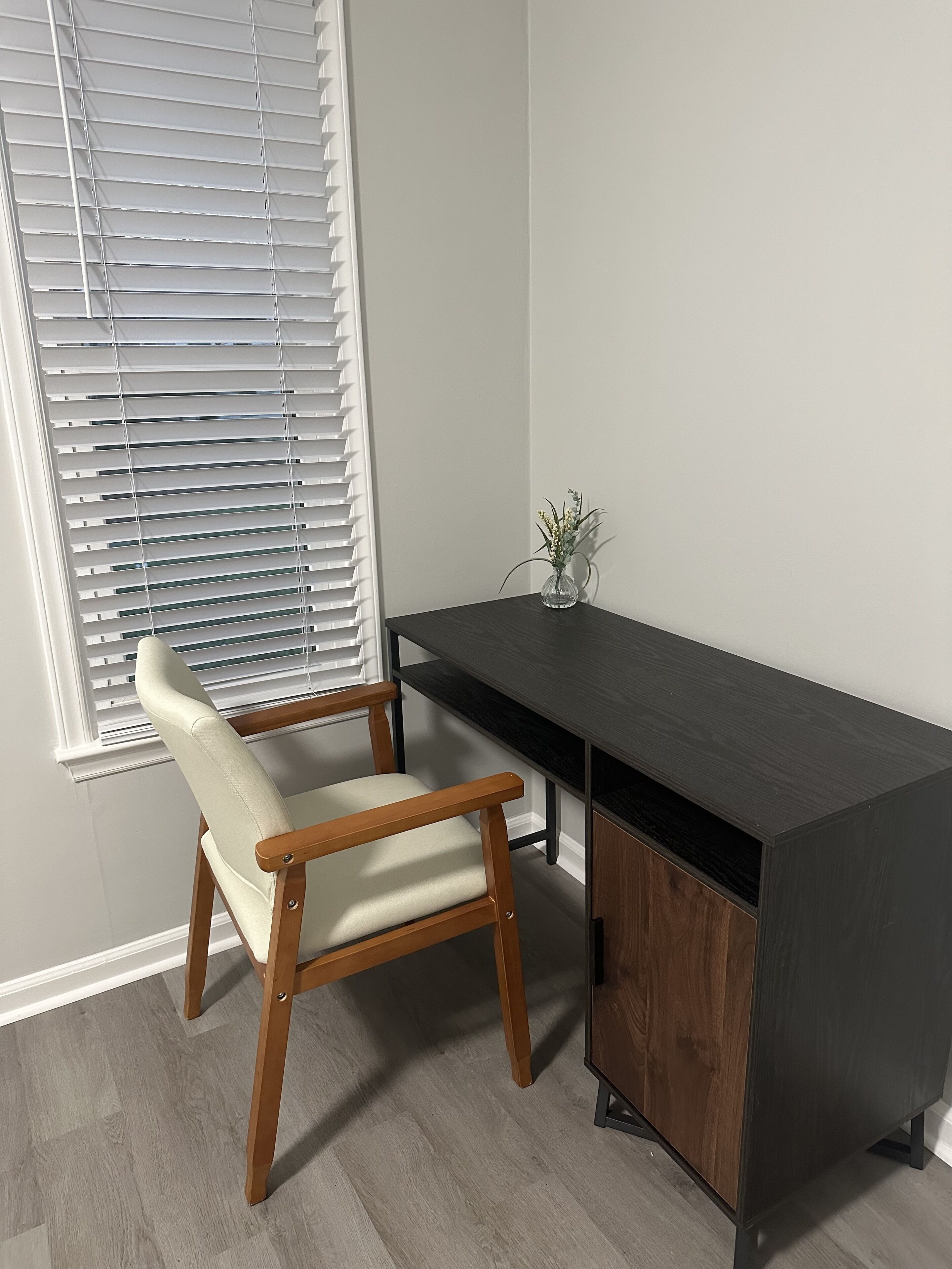 A corner desk with a chair, a small vase with flowers, and a window with blinds.