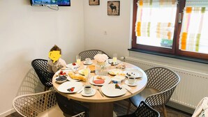 A dining area with a round table set for breakfast, surrounded by wicker chairs. A television is mounted on the wall displaying a scenic view. - Ferienwohnung Busch (Rust)