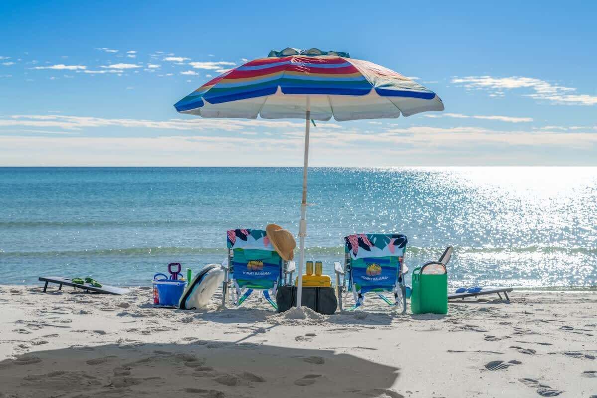 A beach scene with a striped umbrella, two beach chairs, a surfboard, a bucket, and a cooler.
