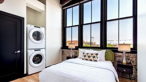 Un dormitorio con cama, lavadora, ventana con vista a la ciudad y pared de ladrillo.