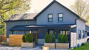 Una casa moderna de dos pisos con fachada gris, un porche con macetas de madera y un pequeño pozo de fuego circular.