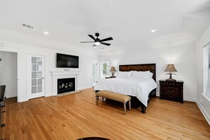 Ein geräumiges Schlafzimmer mit Holzdielenboden, einem großen Bett, einem Kamin, einem Deckenventilator und einem an der Wand montierten Fernseher.