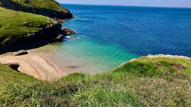 Ett kustlandskap med fri utsikt över havet, grönskande kullar och en sandstrand.