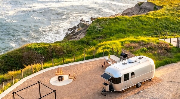 A camper parked on a curved path with a view of the ocean. There is a small seating area with a table and chairs.