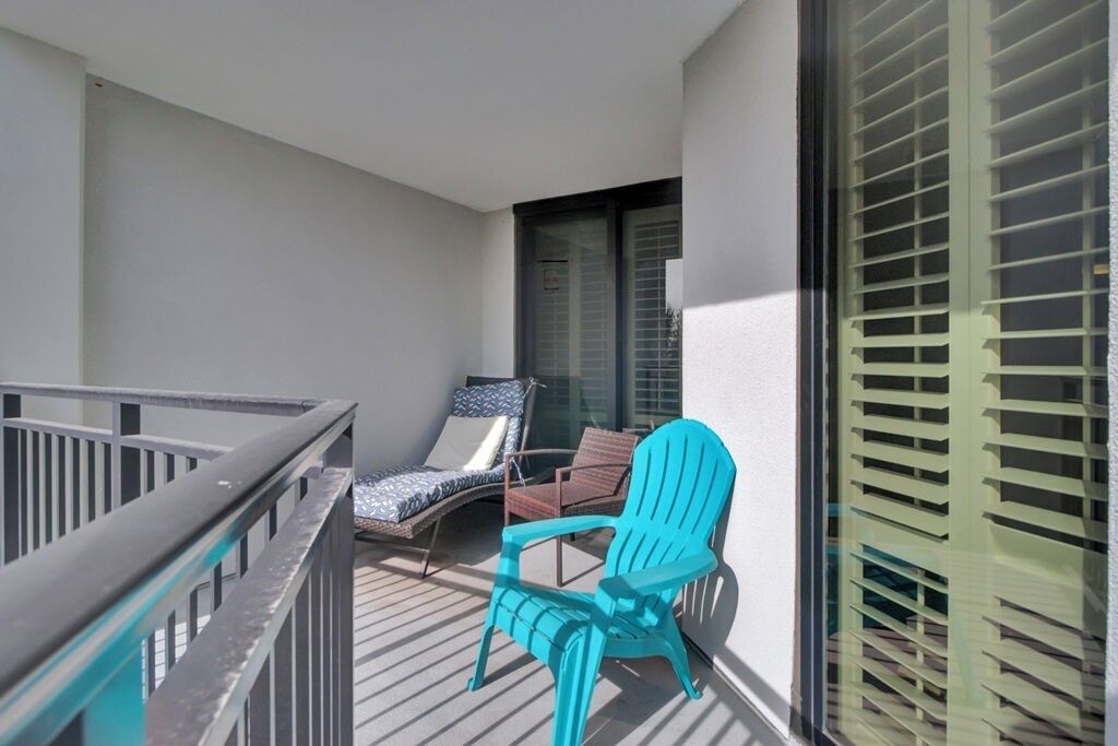 A balcony with a view of the building's exterior, a blue Adirondack chair, a brown wicker chair, and a glass door.