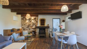 Una sala moderna con chimenea de piedra, vigas de madera en el techo, un comedor con sillas blancas y un televisor de pantalla plana montado en la pared.