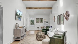 Een ruime hal met houten plafond, een witte bank, een groene fauteuil en een eetgedeelte met tafel en stoelen.