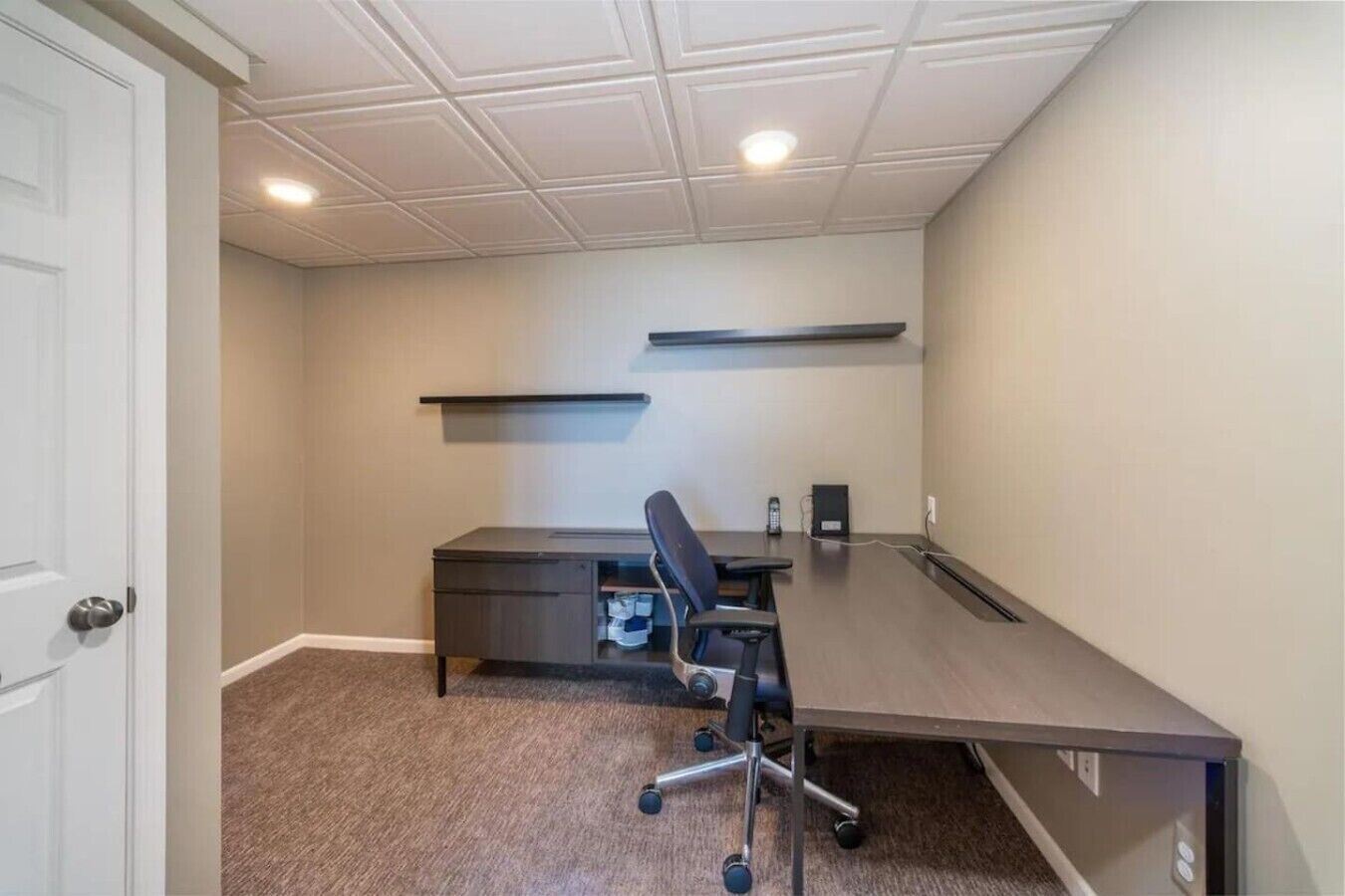 A room with a dark wooden desk, a black office chair, a telephone, and a shelf on the wall.