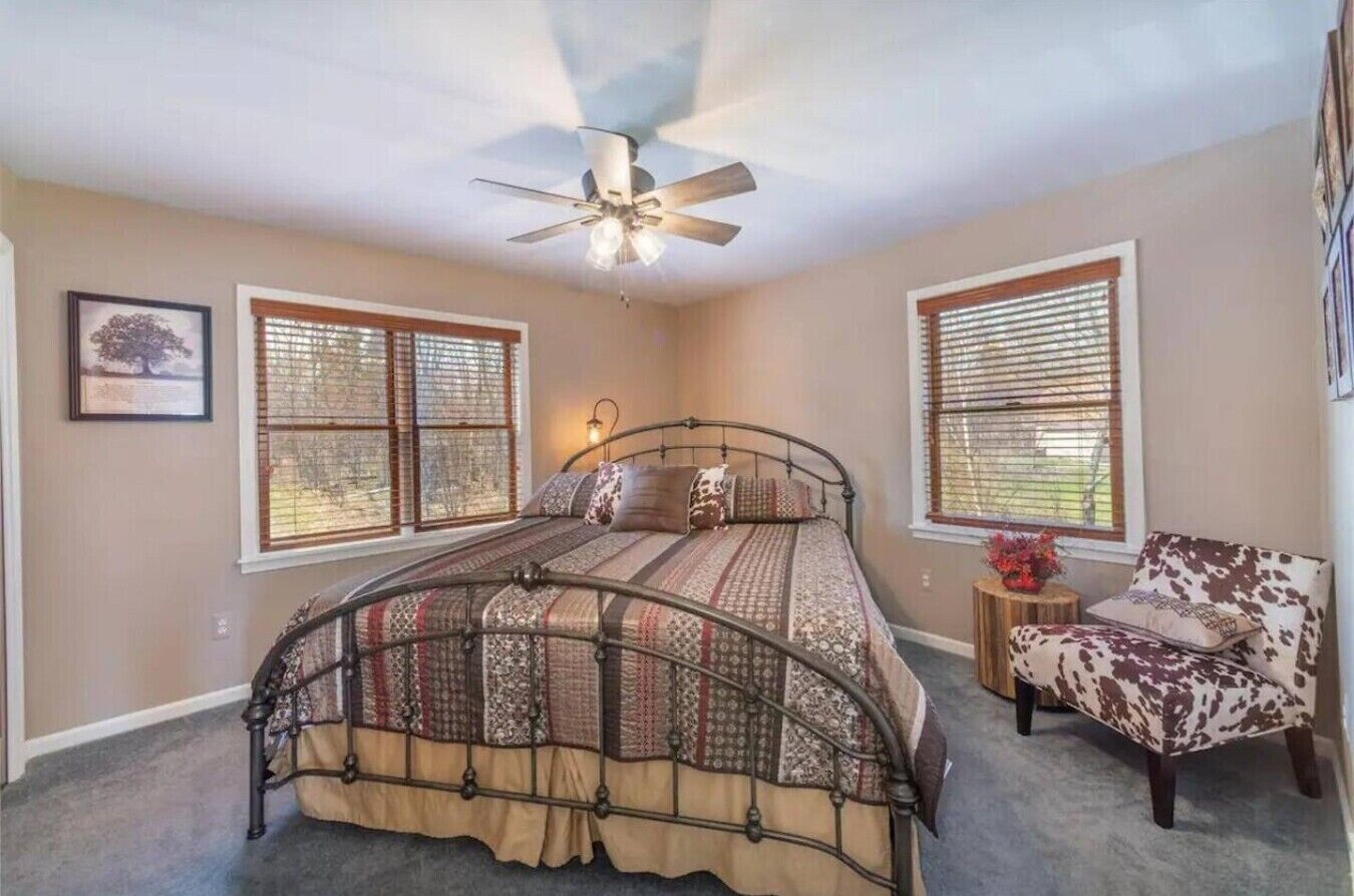 Un dormitorio con cama, una silla, ventilador de techo y dos ventanas con persianas.