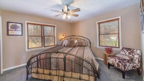 Une chambre à coucher comprenant un lit, une chaise, un ventilateur de plafond et deux fenêtres équipées de stores.