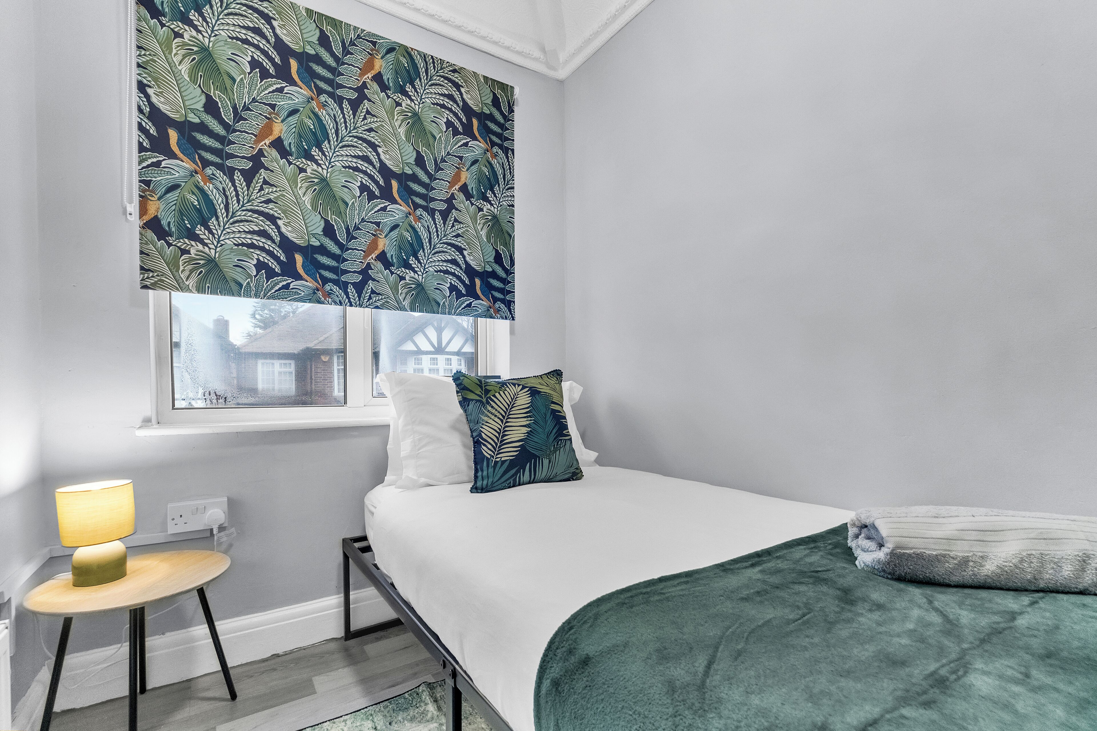 A bedroom with a bed, a window with a leaf-patterned blind, a bedside table with a lamp, and a green throw blanket.