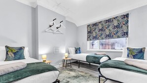 Un dormitorio moderno con dos camas individuales, una mesita de noche pequeña con una lámpara, un radiador y una ventana con persiana estampada con motivos de hojas tropicales.