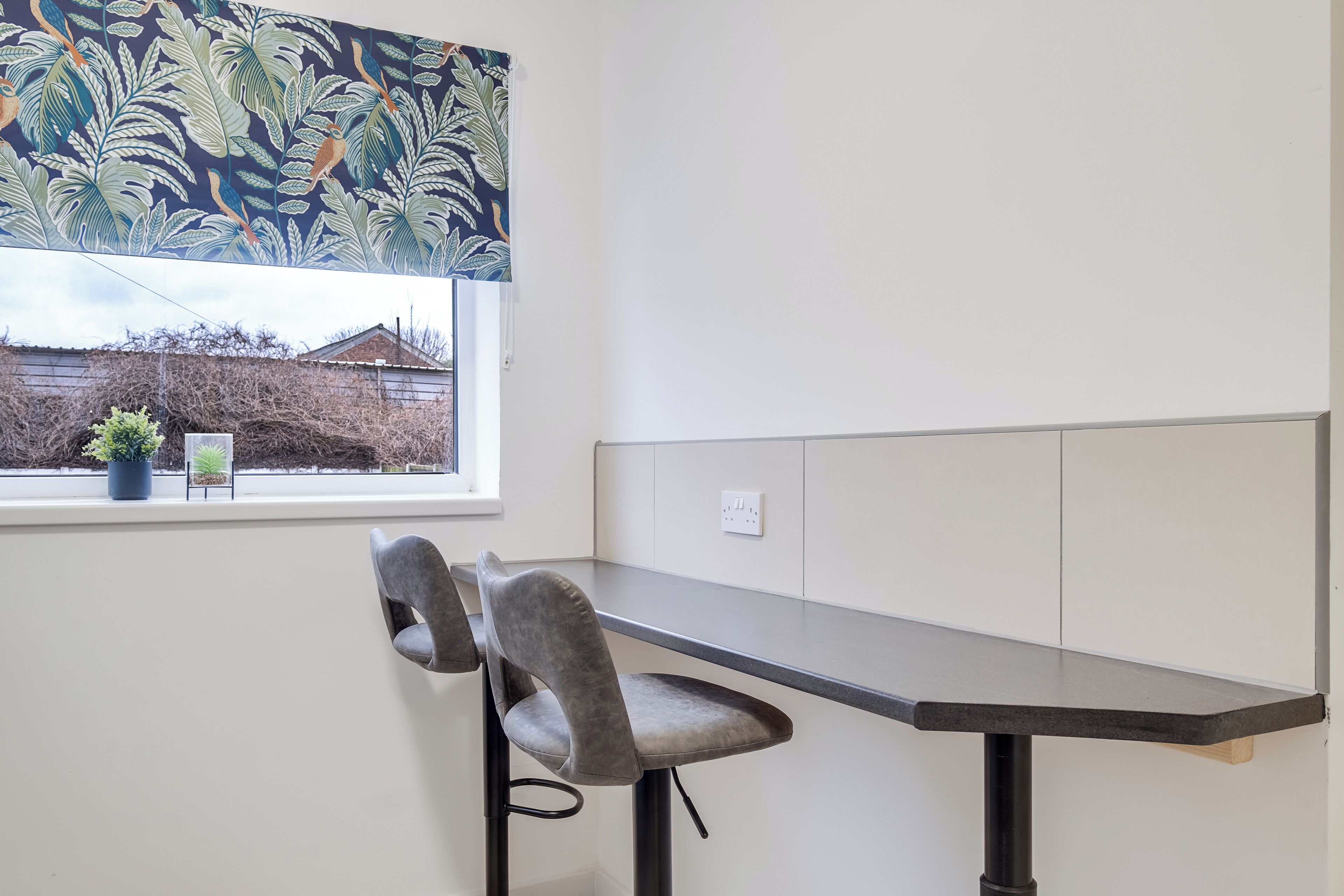 Een hoekbureau met een bureau en twee stoelen, een raam met uitzicht op een huis en een bloempot op de vensterbank.