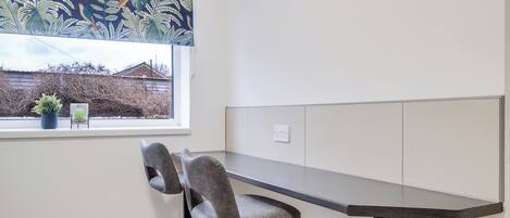 Een hoekbureau met een bureau en twee stoelen, een raam met uitzicht op een huis en een bloempot op de vensterbank.