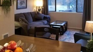 Un salon avec un canapé, une table à manger ornée de fruits, un ventilateur de plafond et une fenêtre donnant sur des arbres.