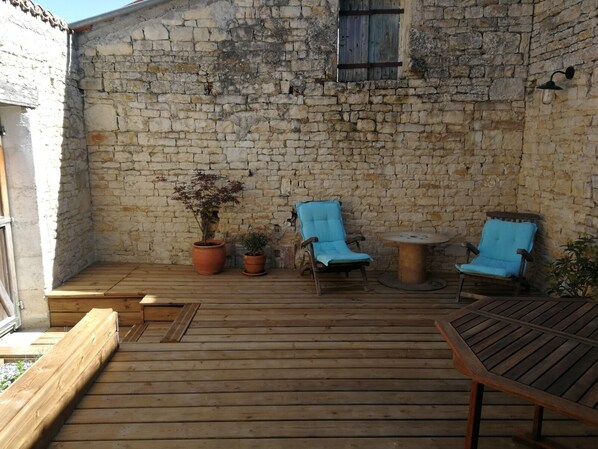 A rustic outdoor patio with wooden flooring, two blue cushioned chairs, a small round table, and potted plants.