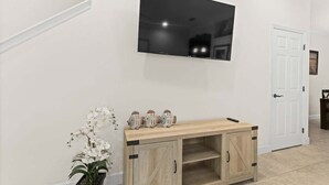 Uma sala de estar moderna com uma TV de tela plana fixada na parede, uma mesinha de madeira com objetos decorativos e uma orquídea em vaso.