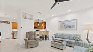 Uma sala de estar moderna com sofá, poltrona e banqueta, uma área de jantar com mesa e cadeiras, e uma cozinha ao fundo.