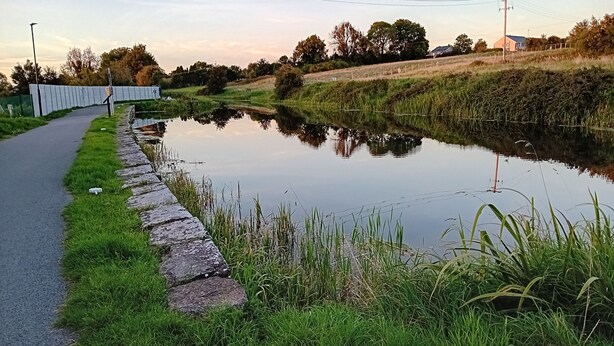 Un canal tranquilo con un muro de piedra y césped a un lado, y un camino pavimentado que lo recorre.