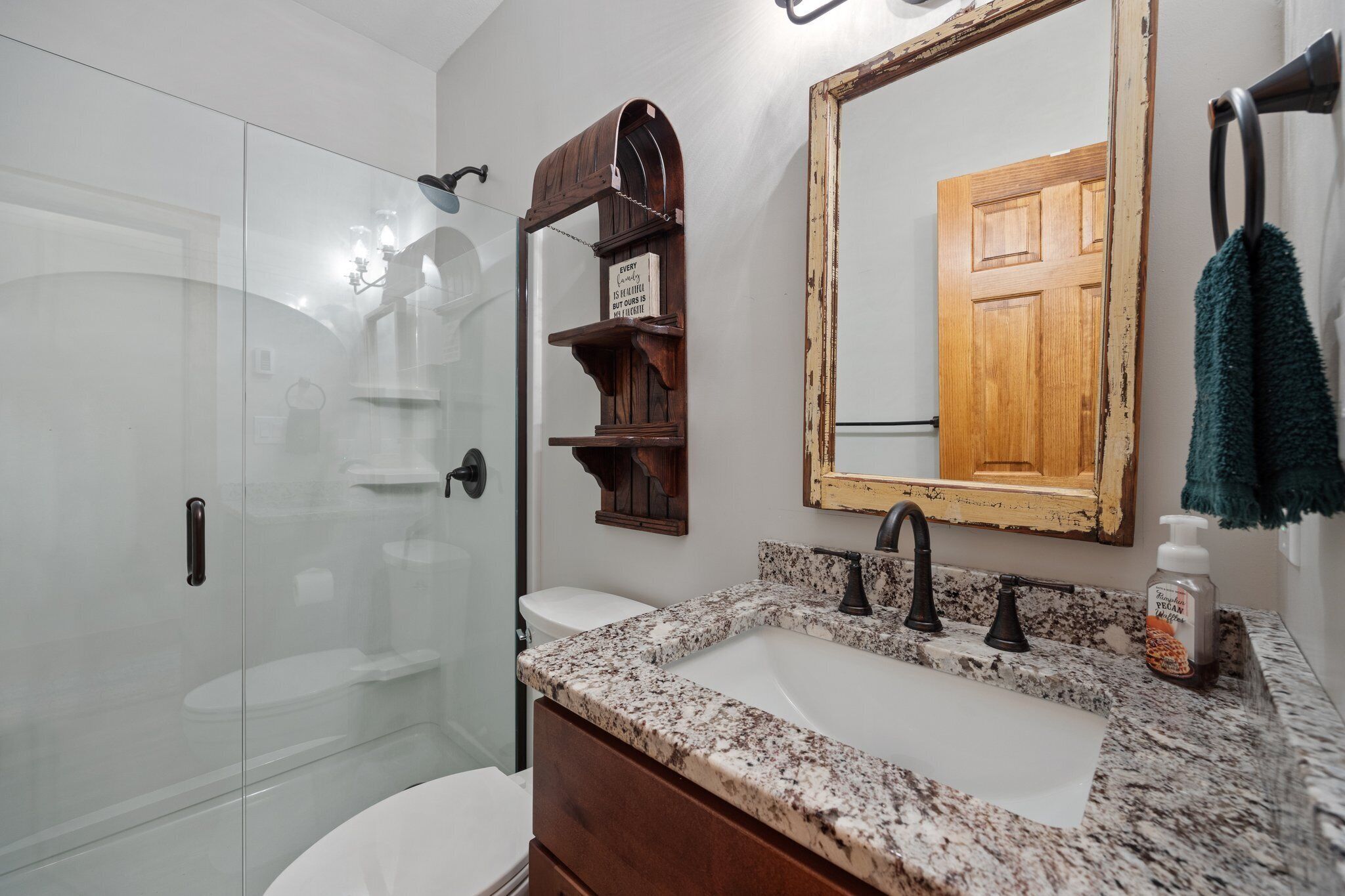 A bathroom with a glass shower enclosure, a wooden shelf with a sign, a large mirror, and a granite countertop.