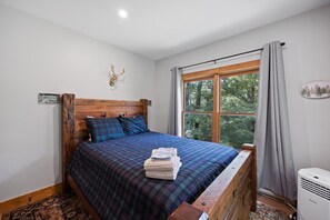 Un dormitorio con cabecera de madera, colcha a cuadros, ventana con vistas a árboles y cabeza de ciervo montada en la pared.