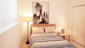 Une chambre d’hôtel avec un lit, deux lampes de chevet, une œuvre d’art représentant un paysage urbain accrochée au mur, et une fenêtre avec des rideaux légers.