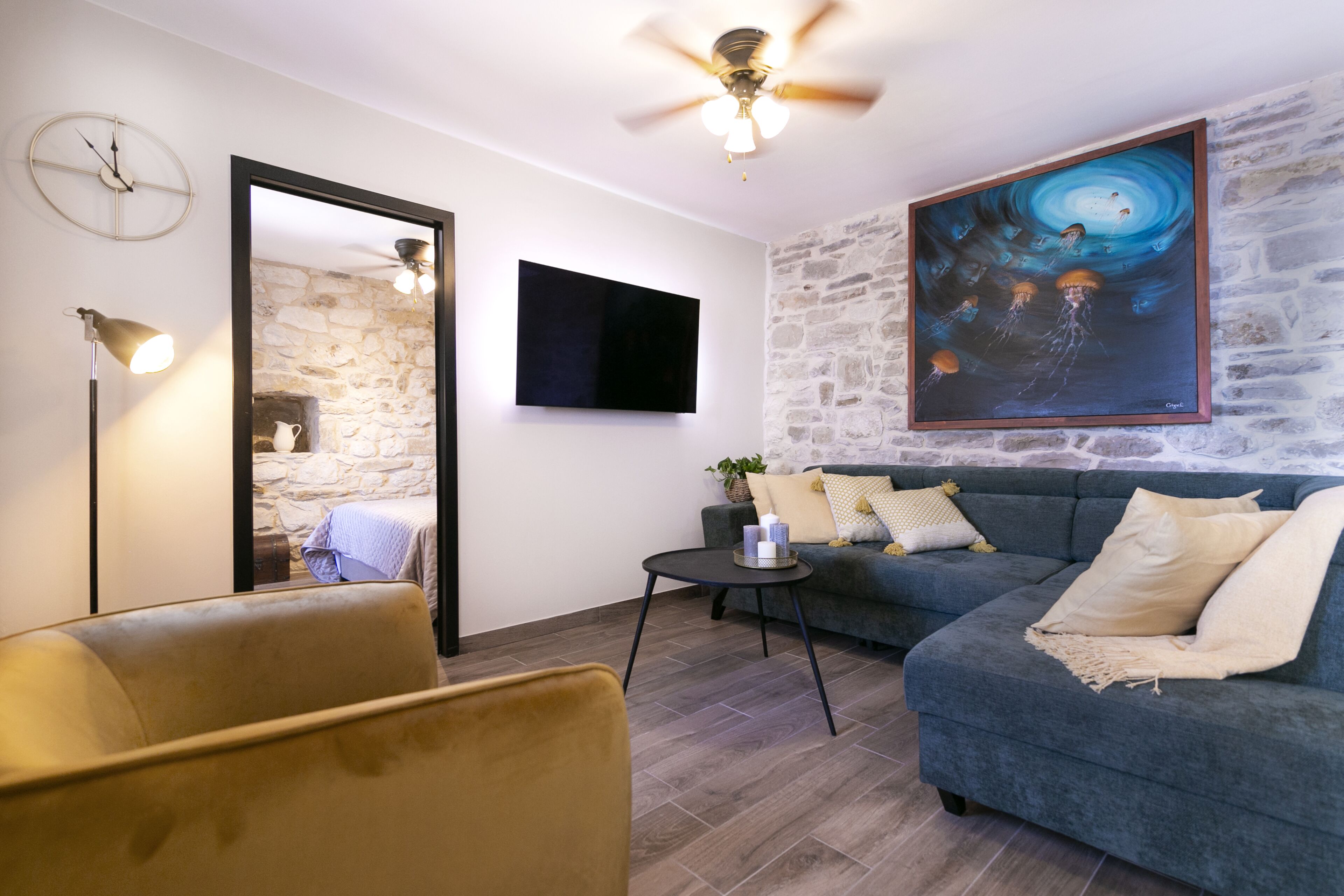 A modern living room with a stone wall, a blue sofa, a yellow armchair, a black coffee table, a ceiling fan, a mounted TV, and a framed painting.