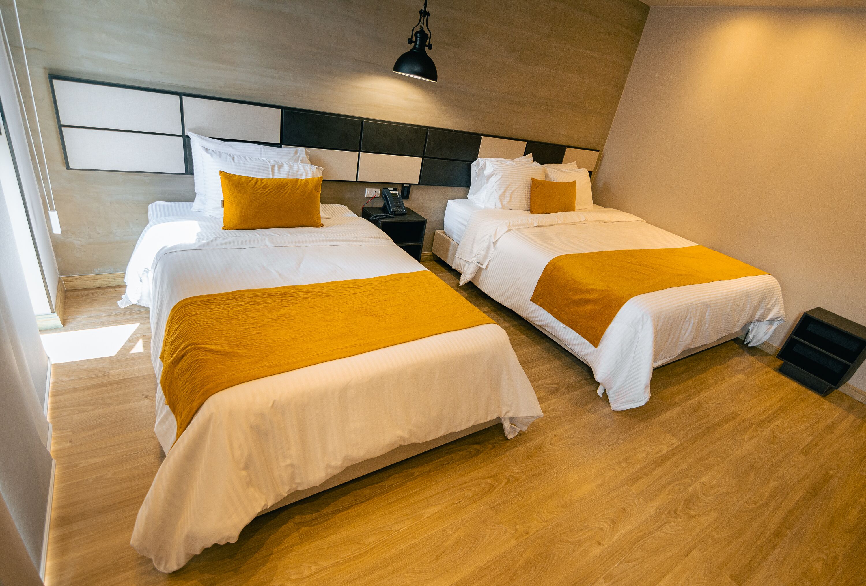 Une chambre d’hôtel avec deux lits, un plancher en bois, une tête de lit moderne et une table de chevet avec une lampe.