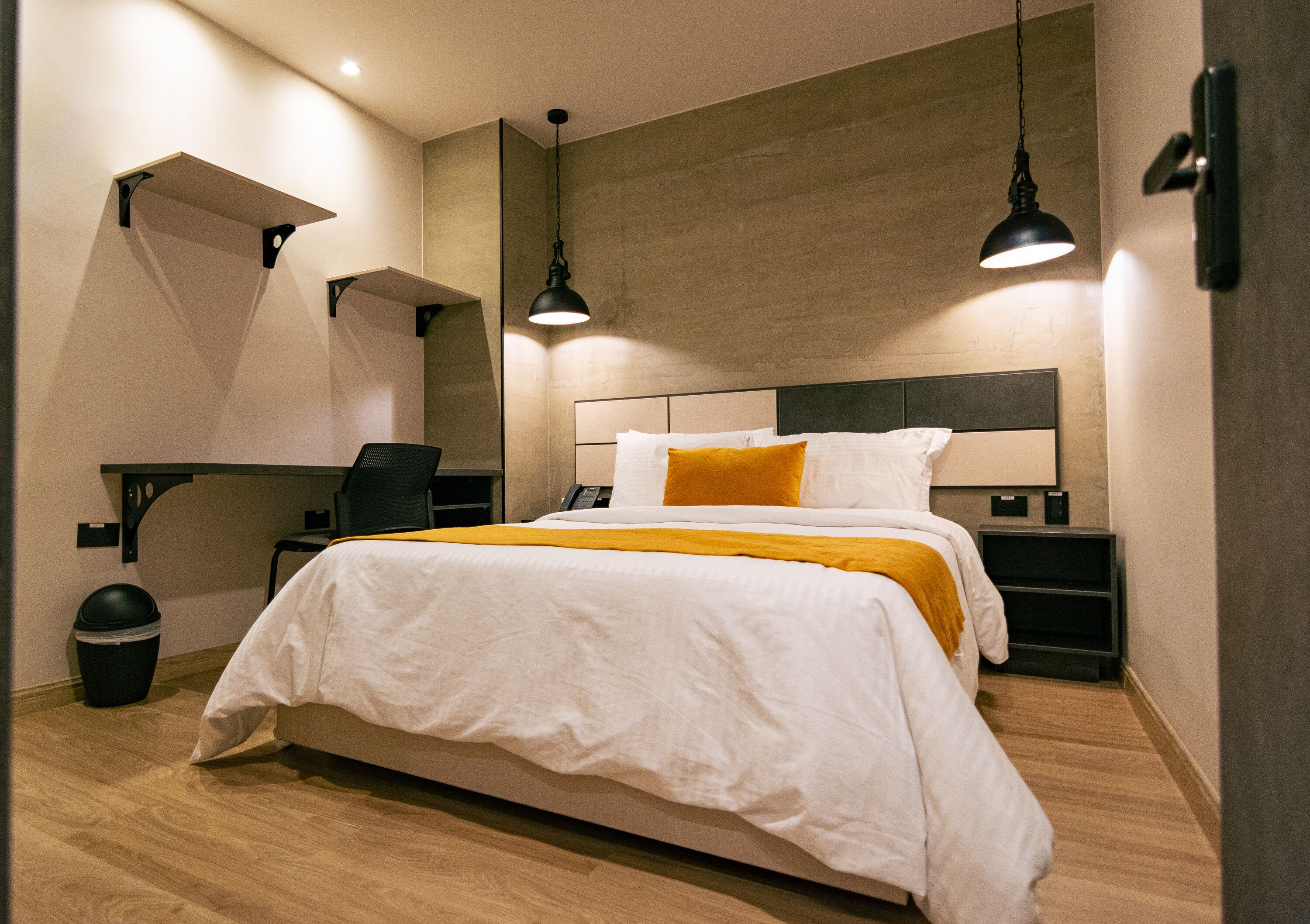 A modern hotel room with a large bed, a desk, and a chair. There is a trash can and a wall-mounted shelf.