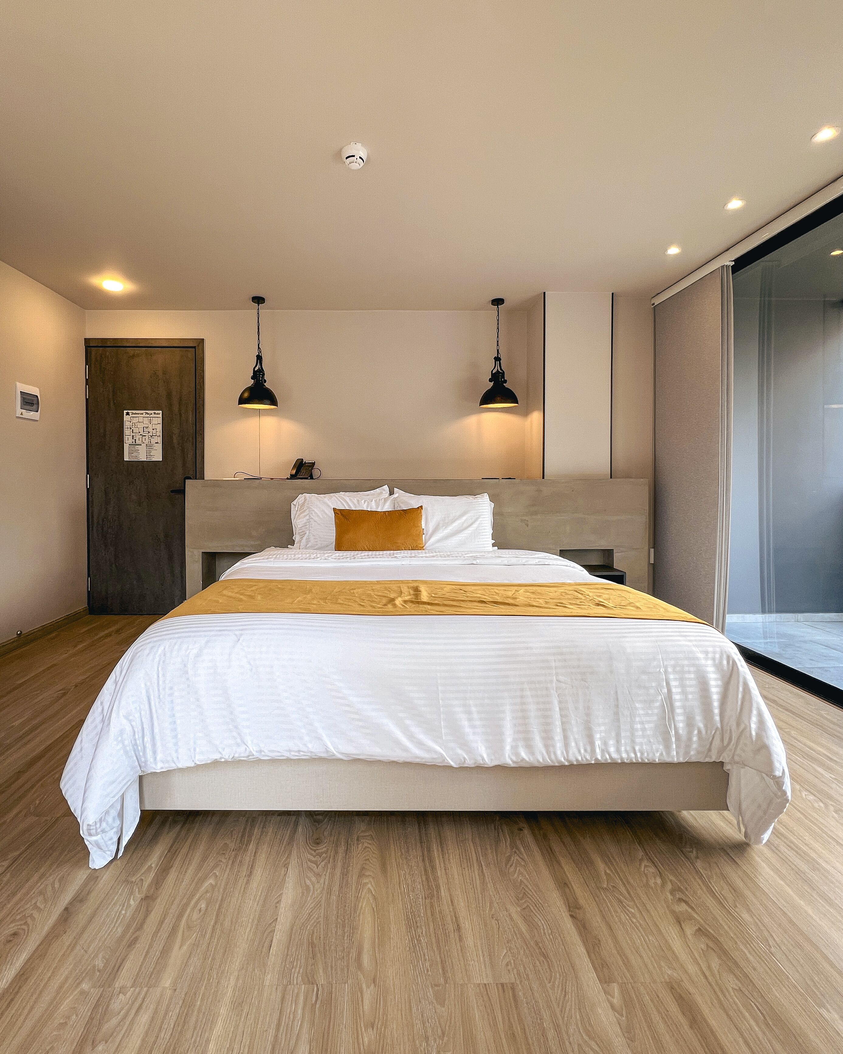 A modern hotel room with a large bed, wooden flooring, two pendant lights, a door with a map, and a large window.